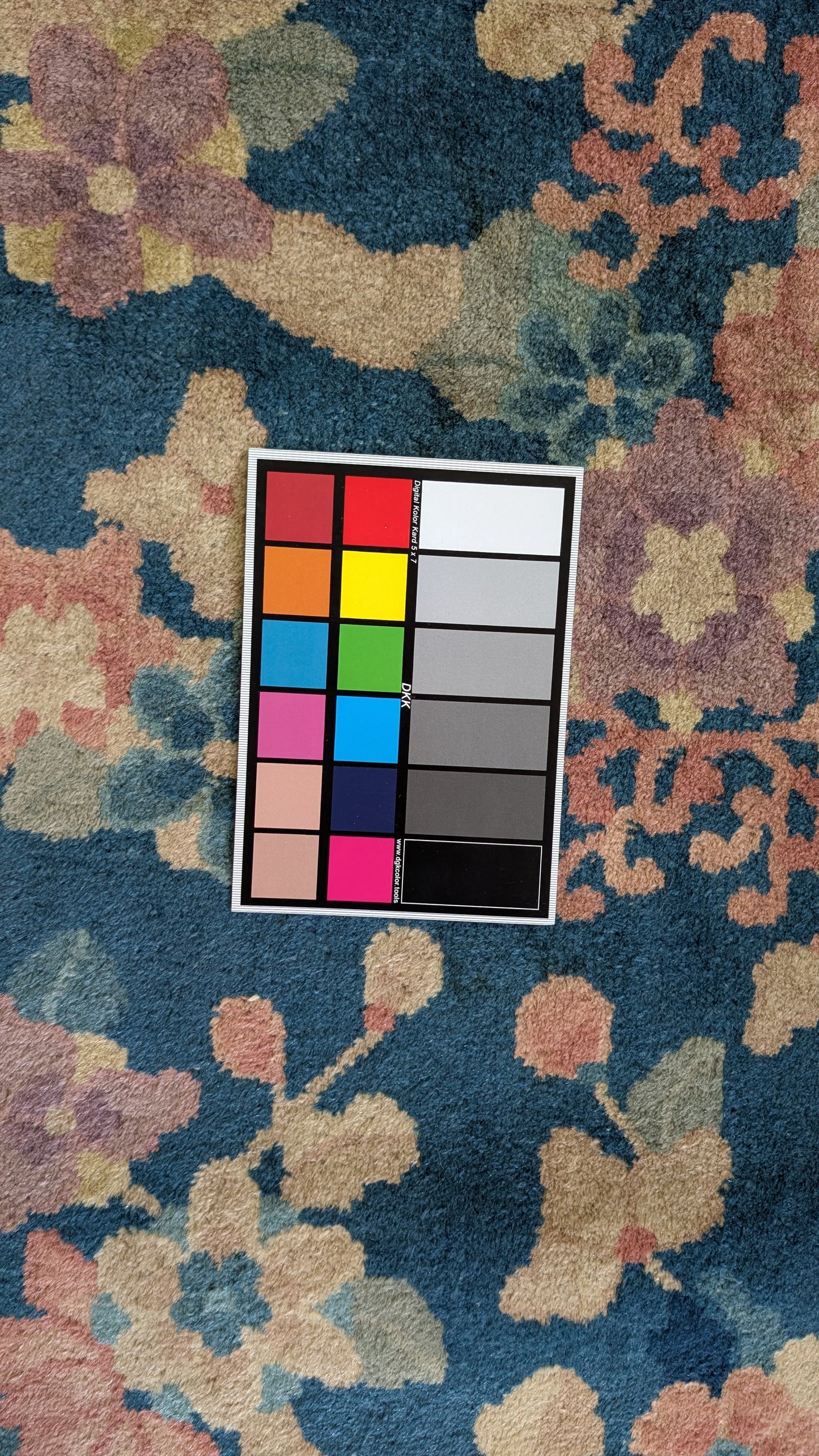 A square color calibration card with multiple color blocks and gray/white reference tiles is placed on top of a plush floral area rug. The rug has a deep blue base with large abstract flowers and leaves in muted pink, beige, and green tones.